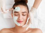 Beautician applying clay face mask on woman face, top view