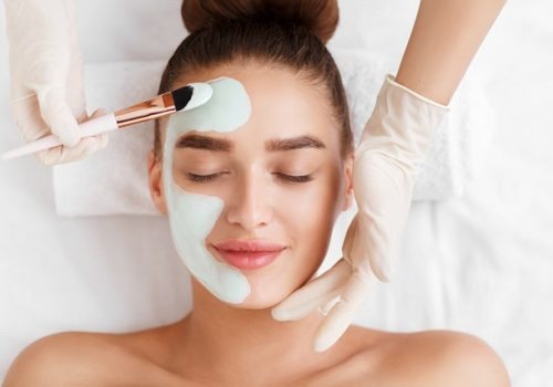 Beautician applying clay face mask on woman face, top view